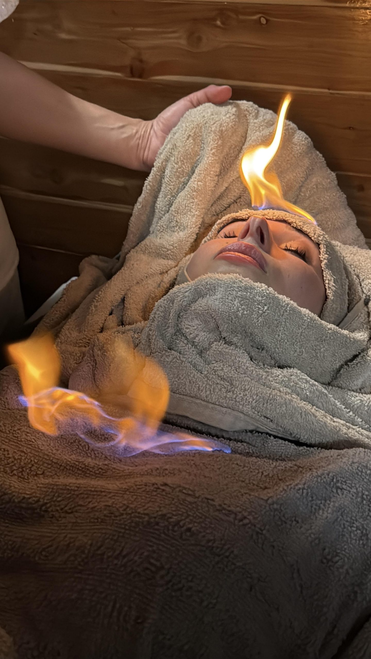 ### Main Subjects - Woman receiving a treatment - Person administering the treatment### Objects - Towels wrapped around the woman - Flames visible near the woman's head### Notable Features - Fire burning gently above the woman's forehead - Relaxed expression on the woman's face - Wooden interior suggesting a spa or wellness environment### Text or Symbols - No text or symbols present### Overall Mood or Theme The scene conveys a unique blend of relaxation and tension, highlighting a spa treatment that incorporates fire. The woman's serene expression contrasts with the flames, creating an intriguing atmosphere that suggests both rejuvenation and risk.