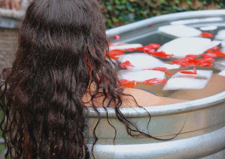 ### Main Subjects - Individual with long, curly hair - Bathtub or tub filled with water### Objects - Ice cubes in the water - Rose petals scattered on the surface - Natural greenery in the background### Notable Features - The tub appears metallic and rustic - The setting is outdoors, surrounded by lush greenery### Text or Symbols - No visible text or symbols present### Overall Mood or Theme The image conveys a serene and tranquil atmosphere, suggesting relaxation and self-care. The combination of the ice, water, and rose petals creates a calming and refreshing aesthetic, inviting a sense of peace and rejuvenation.
