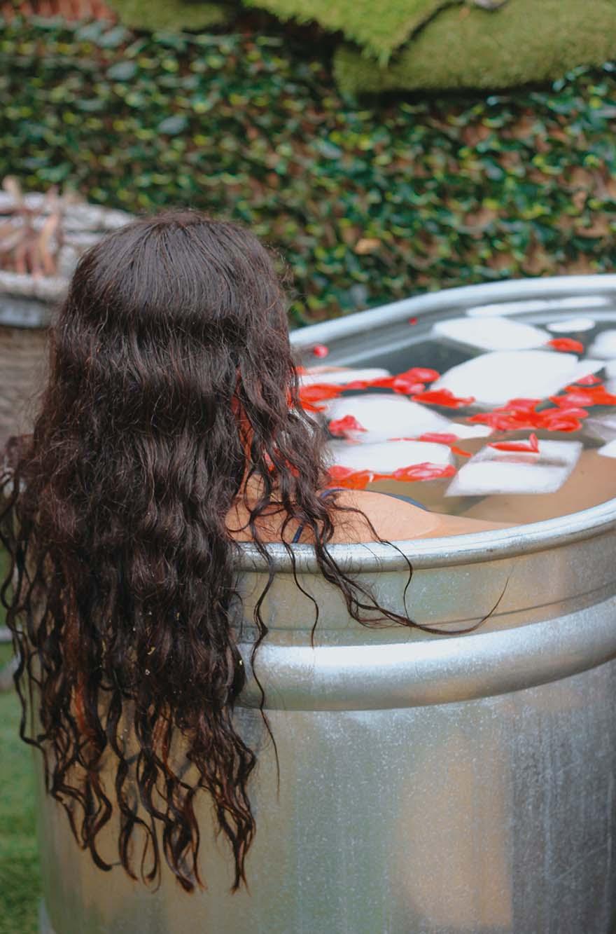 ### Main Subjects - Individual with long, curly hair - Bathtub or tub filled with water### Objects - Ice cubes in the water - Rose petals scattered on the surface - Natural greenery in the background### Notable Features - The tub appears metallic and rustic - The setting is outdoors, surrounded by lush greenery### Text or Symbols - No visible text or symbols present### Overall Mood or Theme The image conveys a serene and tranquil atmosphere, suggesting relaxation and self-care. The combination of the ice, water, and rose petals creates a calming and refreshing aesthetic, inviting a sense of peace and rejuvenation.