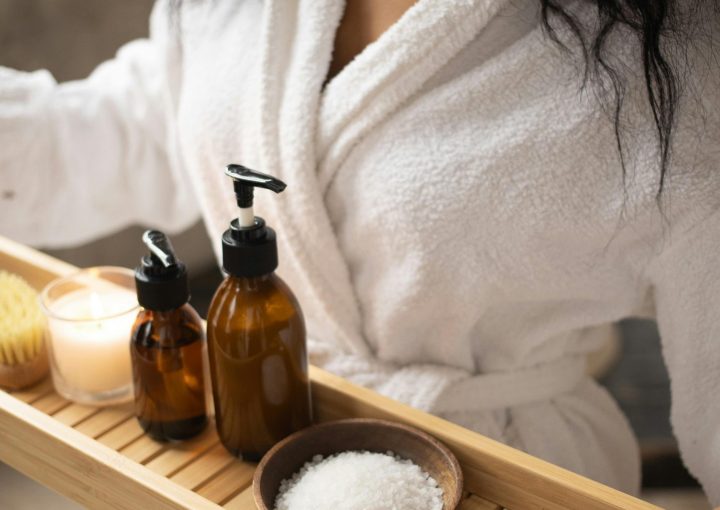 ### Key Elements- **Main Subject**: A woman in a white bathrobe. - **Objects**: A wooden tray holding two amber bottles, a bowl of salt, a candle, and a bath brush. - **Notable Features**: The warm tones of the amber bottles, the texture of the bathrobe, and the calming ambiance created by the candlelight. - **Text/Symbols**: None present.### Overall Mood/Theme The image conveys a serene and relaxing atmosphere, suggesting a spa-like experience focused on self-care and wellness. The soft lighting and natural materials enhance the tranquil theme.